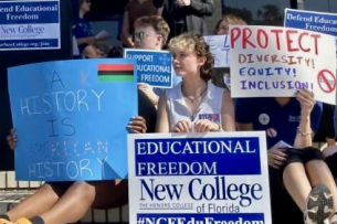 A group of protesters sitting on steps with signs. One sign reads "Protect Diversity Equity Inclusion."